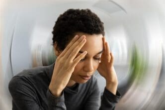 A person with short, curly hair holding their temples and closing their eyes, with a blurred, swirling background indicating dizziness or discomfort.