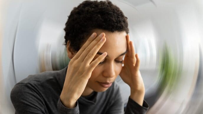 A person with short, curly hair holding their temples and closing their eyes, with a blurred, swirling background indicating dizziness or discomfort.