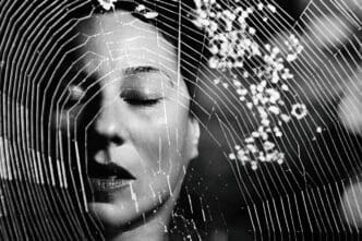 A black and white close-up of a person's face with closed eyes, overlaid by a prominent spider web in sharp focus across the foreground, symbolizing phobia or exposure therapy.