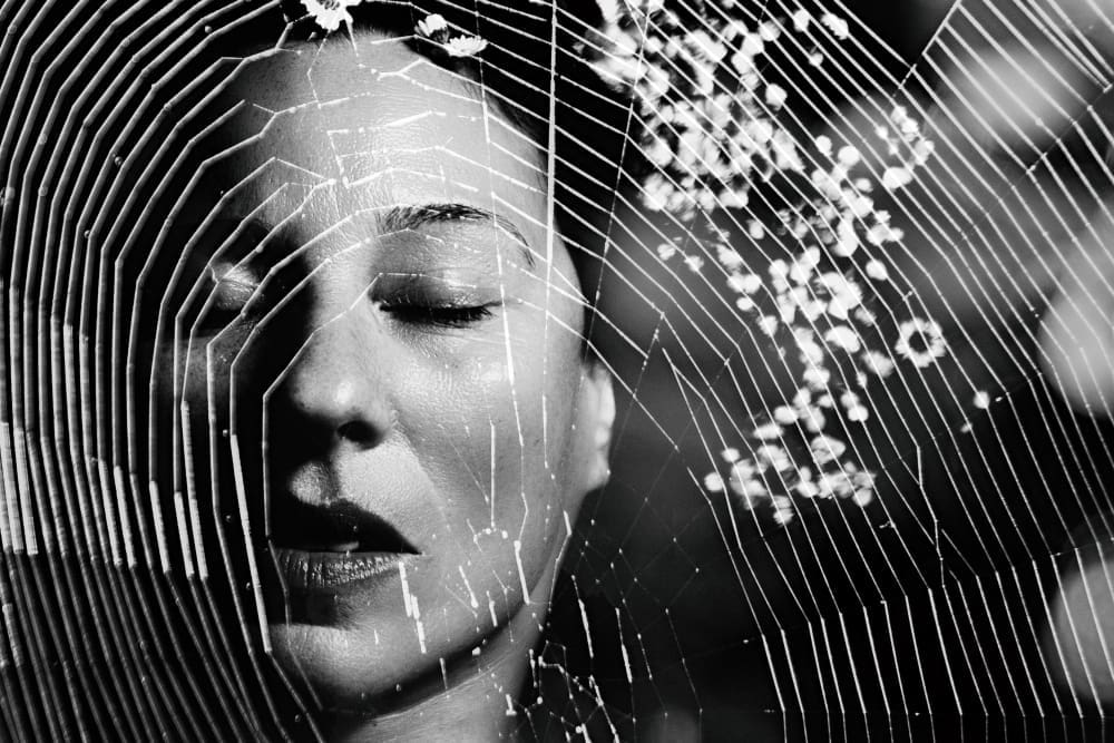 A black and white close-up of a person's face with closed eyes, overlaid by a prominent spider web in sharp focus across the foreground, symbolizing phobia or exposure therapy.