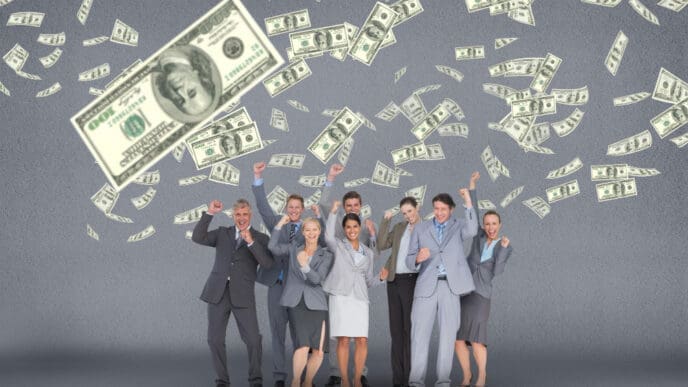 A diverse group of seven business professionals in suits enthusiastically cheer with arms raised as US dollar bills fall around them against a grey background.