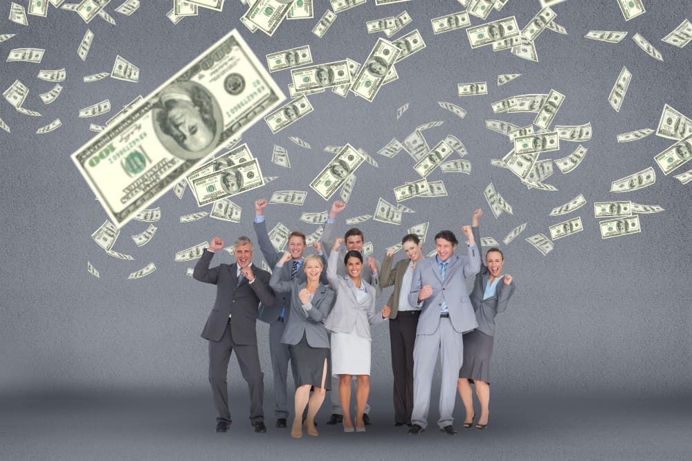 A diverse group of seven business professionals in suits enthusiastically cheer with arms raised as US dollar bills fall around them against a grey background.