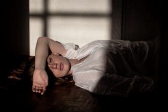 A woman with freckles lying on a dark floor in a beam of sunlight, appearing despondent.