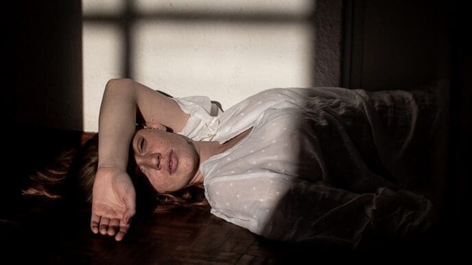 A woman with freckles lying on a dark floor in a beam of sunlight, appearing despondent.