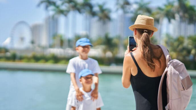A woman in a hat and black top takes a picture with her smartphone of two young boys wearing blue caps and white t-shirts, standing by the water, with palm trees and distant buildings under a clear sky, enjoying a family day in Miami.