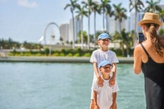 A mother wearing a hat and black top takes a picture with her smartphone of her two young sons, both in white t-shirts and blue caps, standing by a body of water with Miami's Bayside Marketplace and Ferris wheel in the background.