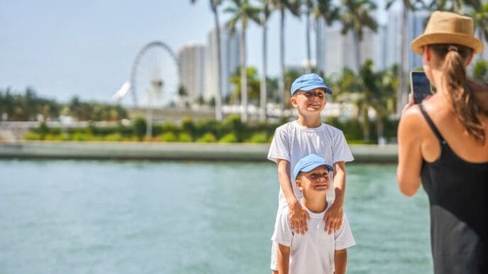 A mother wearing a hat and black top takes a picture with her smartphone of her two young sons, both in white t-shirts and blue caps, standing by a body of water with Miami's Bayside Marketplace and Ferris wheel in the background.