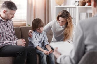 A worried mother comforting her child, who appears sad, while a father and a therapist are present in the room, suggesting a family therapy session.