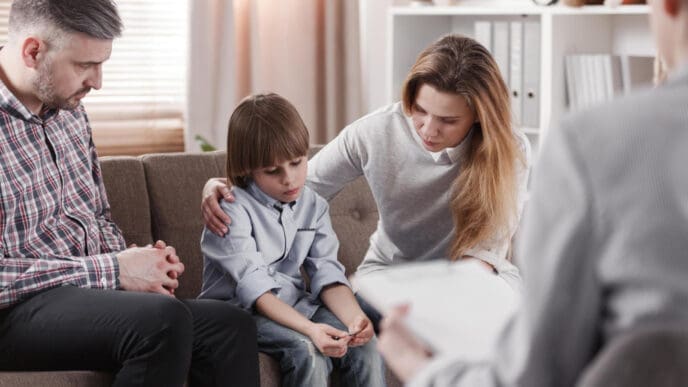 A worried mother comforting her child, who appears sad, while a father and a therapist are present in the room, suggesting a family therapy session.