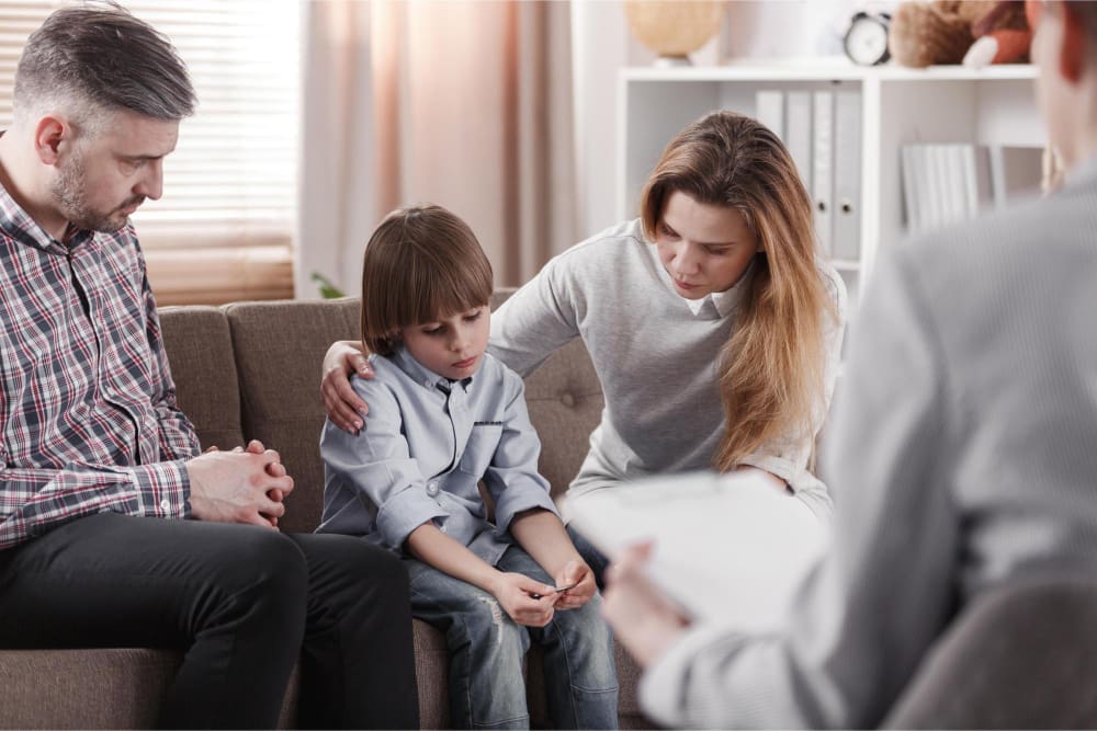 A worried mother comforting her child, who appears sad, while a father and a therapist are present in the room, suggesting a family therapy session.