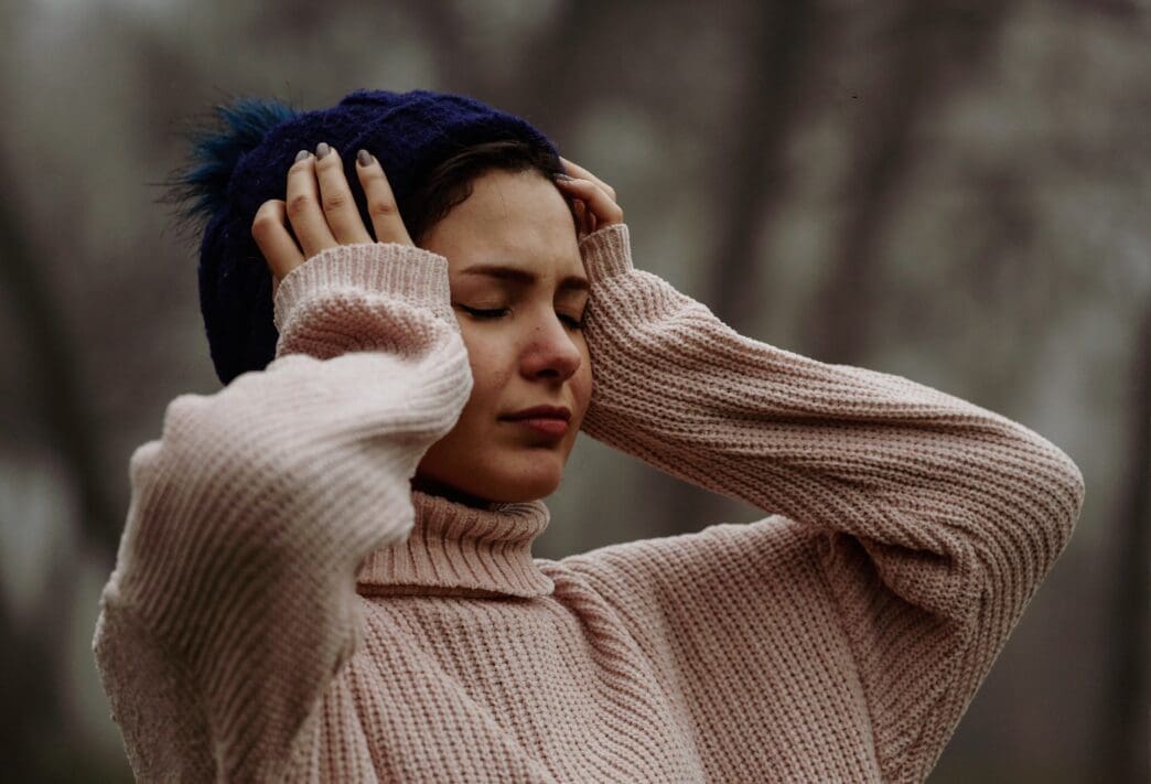 A young woman holding her head, with eyes closed and a pained expression, in a foggy outdoor setting.
