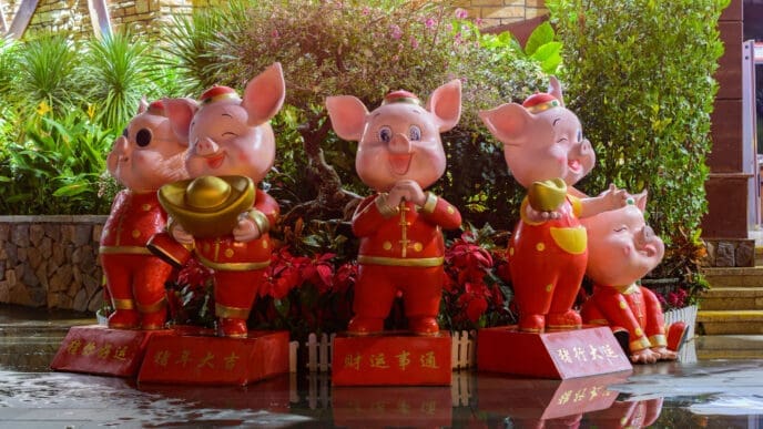Five cheerful pig statues, adorned in traditional red and gold attire, standing on pedestals with Chinese characters, amidst lush green and red foliage, celebrating the Year of the Pig.