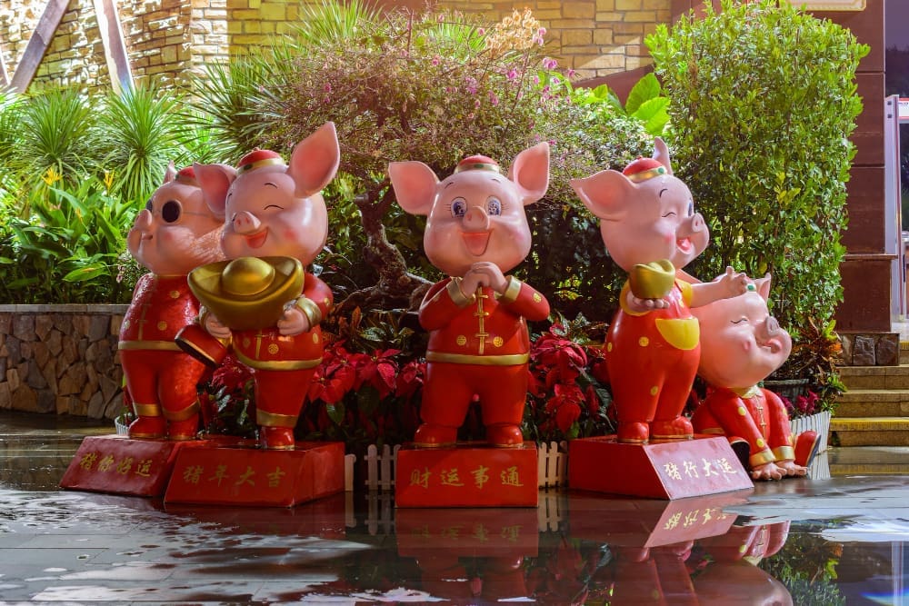 Five cheerful pig statues, adorned in traditional red and gold attire, standing on pedestals with Chinese characters, amidst lush green and red foliage, celebrating the Year of the Pig.