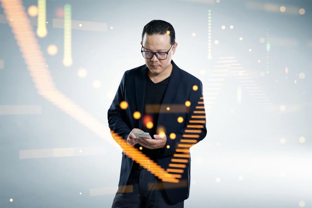 A man in a dark blazer and glasses looks down at his smartphone, surrounded by glowing orange and green digital data streams and abstract light patterns, on a white background.