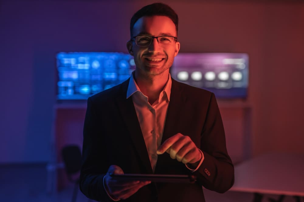 A smiling man in a suit and glasses holds a tablet, looking at the camera, with a background of glowing blue and pink digital screens, suggesting a FinTech or tech-driven environment.