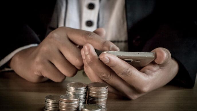 A person's hands are busy on a smartphone, with stacks of silver coins on a wooden desk in the foreground, symbolizing digital finance and investment.