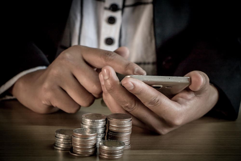 A person's hands are busy on a smartphone, with stacks of silver coins on a wooden desk in the foreground, symbolizing digital finance and investment.
