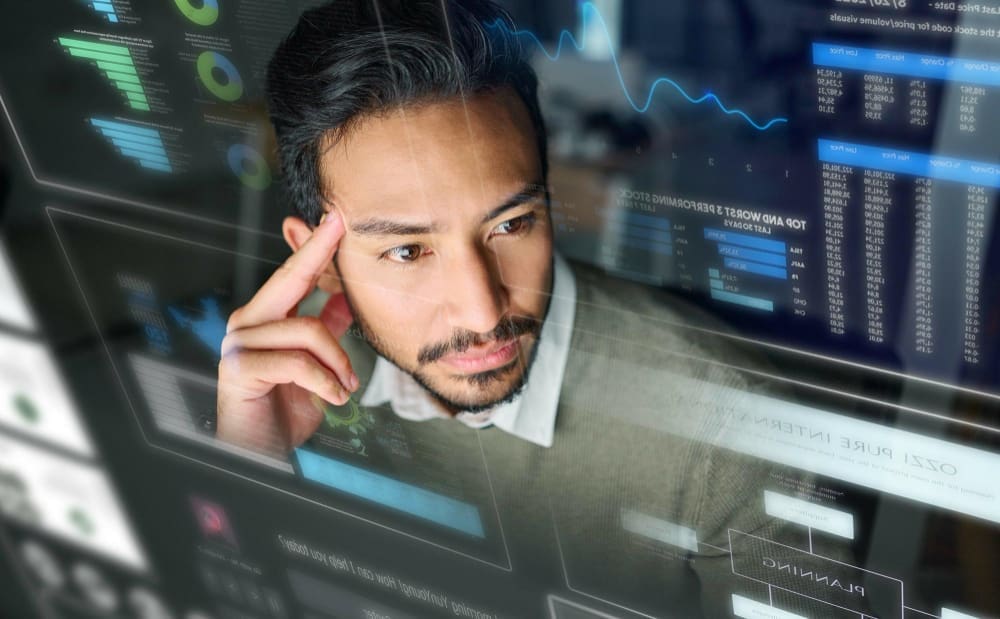 A man with a beard and dark hair, wearing a collared shirt, looks intently at multiple transparent digital screens displaying various graphs, charts, and data, representing complex FinTech tools.