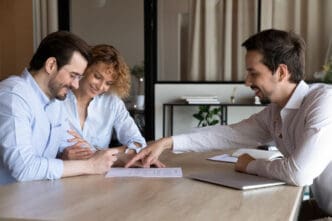 A smiling couple signing a document while a male financial advisor points to a section on the paper during a meeting.