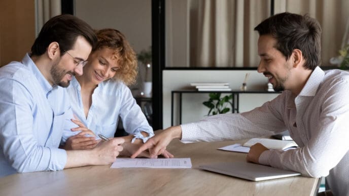 A smiling couple signing a document while a male financial advisor points to a section on the paper during a meeting.