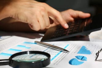A close-up of a person's hands operating a calculator, with financial charts, graphs, a magnifying glass, and a pen on a desk, symbolizing a financial audit.