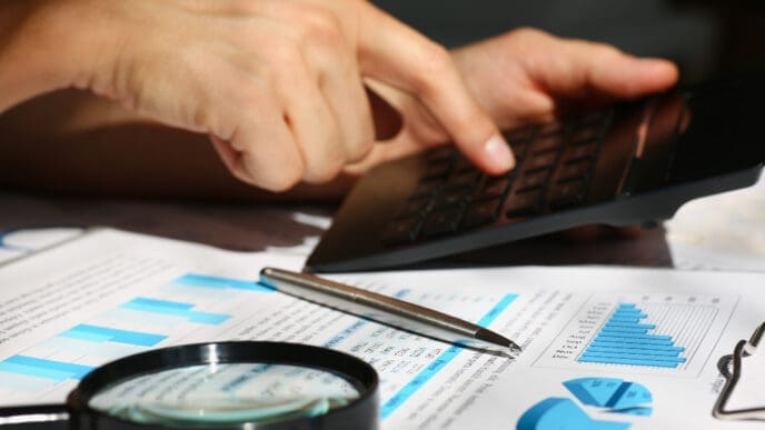 A close-up of a person's hands operating a calculator, with financial charts, graphs, a magnifying glass, and a pen on a desk, symbolizing a financial audit.