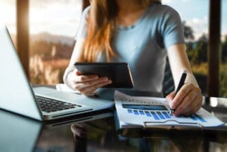 A person working on financial documents with a laptop and tablet, overlooking a scenic view.
