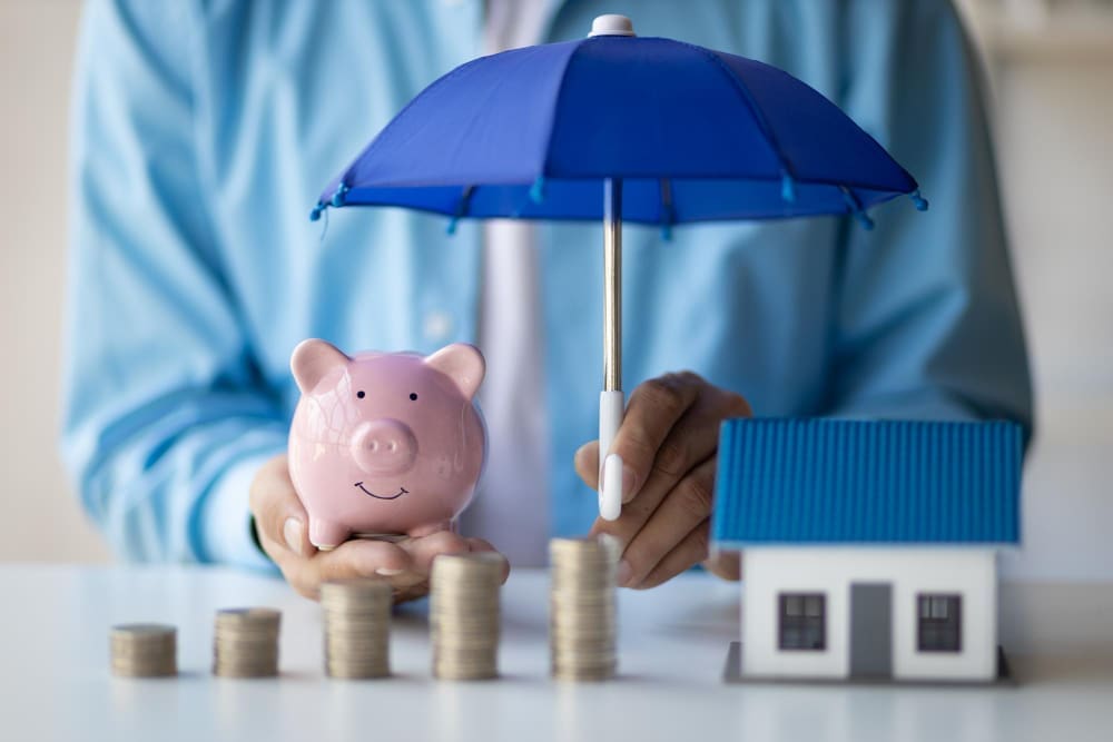 A person holds a blue umbrella over a pink piggy bank and a model house, with stacks of coins arranged in front, symbolizing financial protection and long-term growth.