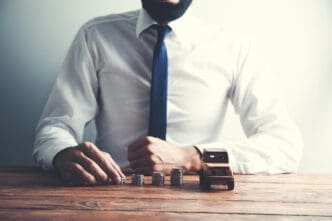 A man in a white shirt and dark tie, with a beard, sits at a wooden table, stacking coins next to a small wooden car, symbolizing financial planning or accumulation of wealth.