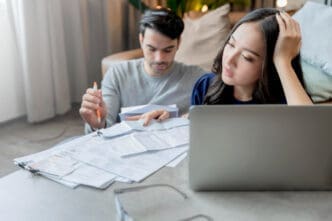 A stressed woman looking at a laptop while a man reviews papers, indicating financial concerns and planning.