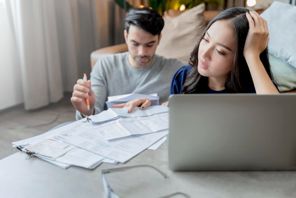 A stressed woman looking at a laptop while a man reviews papers, indicating financial concerns and planning.
