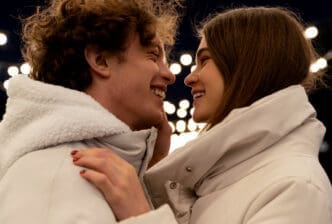 A happy young couple in winter coats looking at each other closely and smiling, with blurred lights in the background.