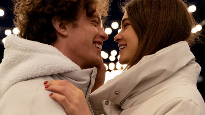 A happy young couple in winter coats looking at each other closely and smiling, with blurred lights in the background.