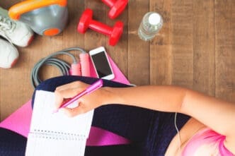 A top-down view of a person writing in a notebook on a pink yoga mat, surrounded by running shoes, red dumbbells, a kettlebell, a jump rope, a water bottle, and a smartphone, symbolizing fitness planning.