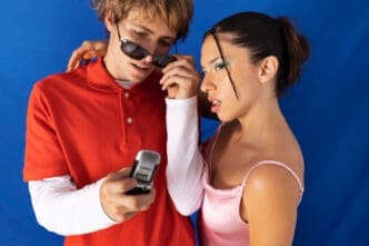 A young man wearing sunglasses and a red polo shirt, looking intently at an old flip phone while a woman in a pink top looks on with interest, against a blue background.