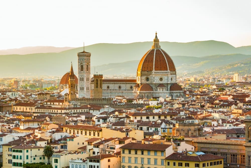 High-angle view of Florence Cathedral (Duomo) and the city skyline at sunset, with mountains in the background.