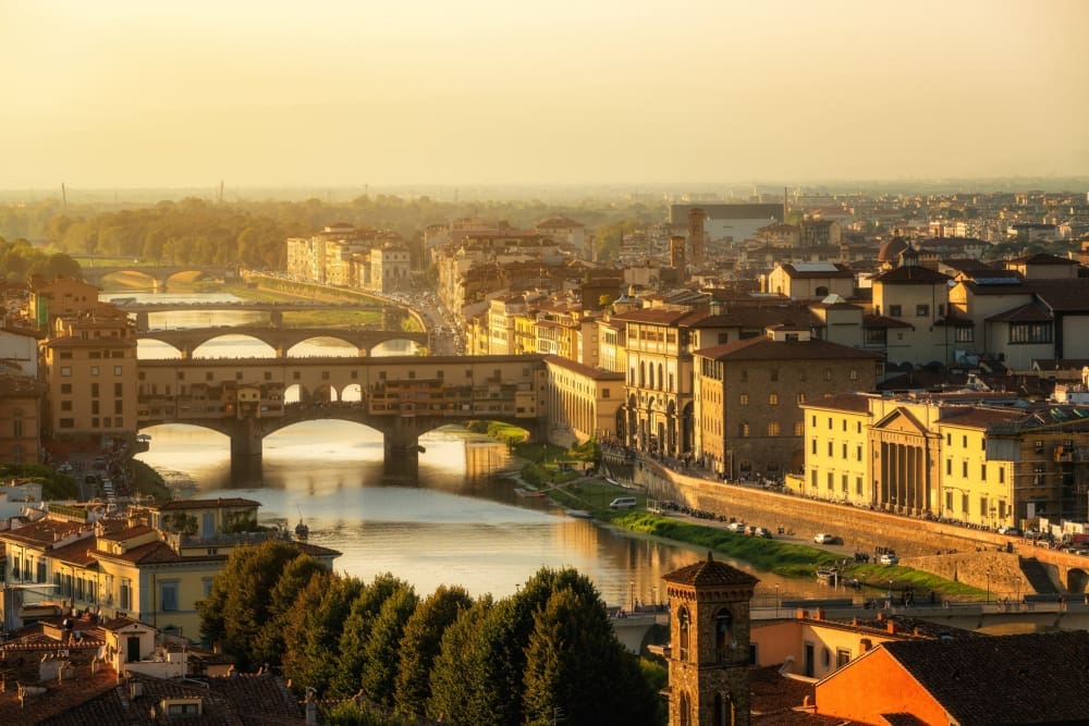 A high-angle view of Florence, Italy, at golden hour, showing the historic Ponte Vecchio bridge with shops, other bridges, and classic buildings lining the Arno River under a hazy, warm-toned sky.