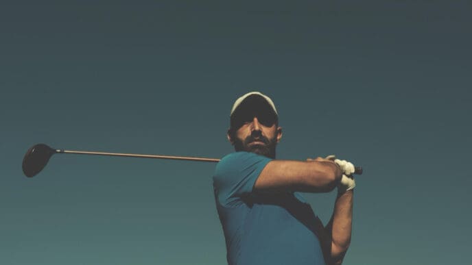 A bearded man in a golf shirt and cap, mid-swing with a golf club, looking directly at the camera under a clear sky.