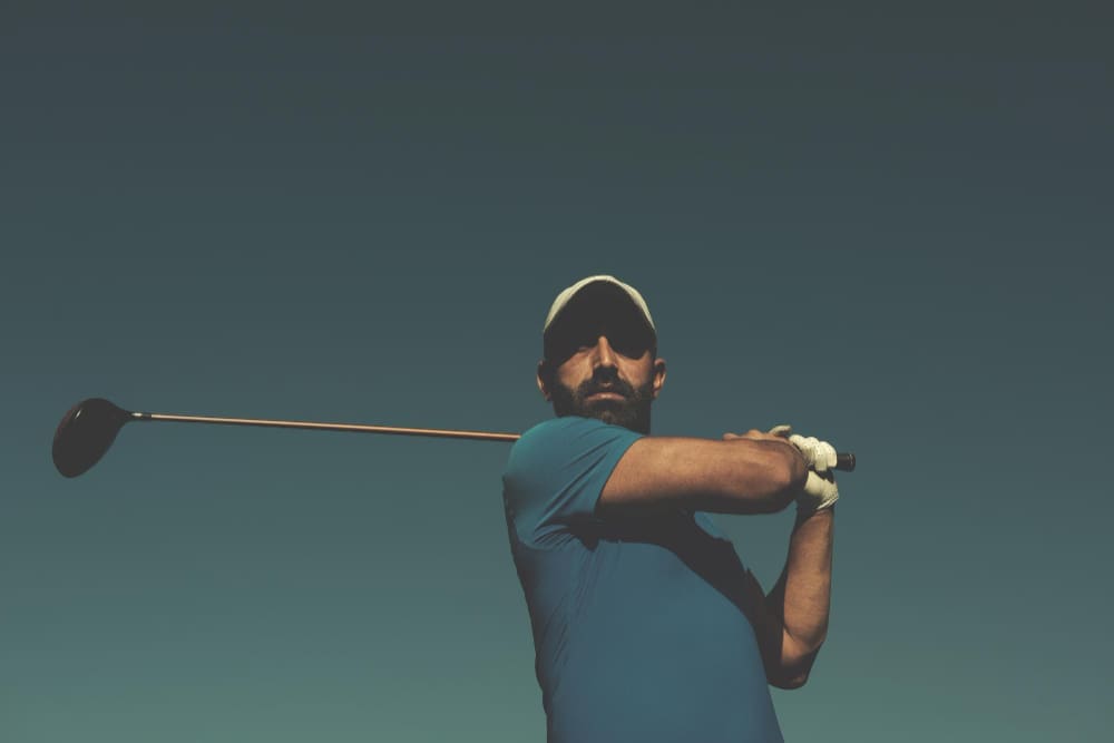 A bearded man in a golf shirt and cap, mid-swing with a golf club, looking directly at the camera under a clear sky.