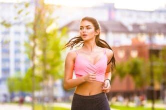 A young woman with long dark hair, wearing a pink sports bra and grey leggings, running outdoors with a determined expression.