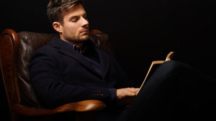 A man with short hair and a beard, wearing a dark coat, sitting in a leather armchair and reading a book in a dimly lit setting.
