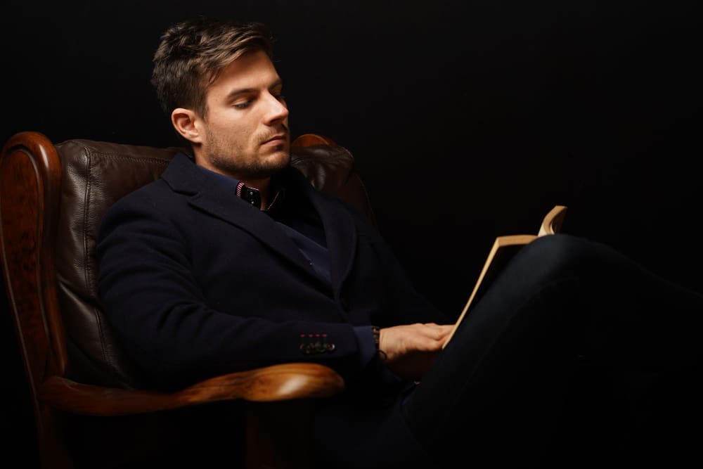 A man with short hair and a beard, wearing a dark coat, sitting in a leather armchair and reading a book in a dimly lit setting.