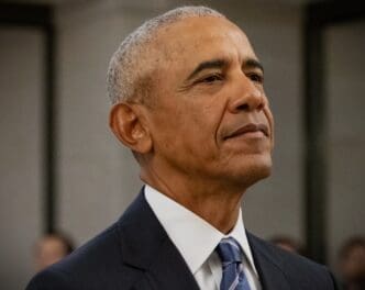 A close-up portrait of former President Barack Obama, looking upwards with a contemplative expression, showing his grey hair and wearing a dark suit with a blue and white striped tie.