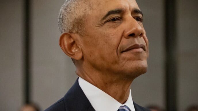 A close-up portrait of former President Barack Obama, looking upwards with a contemplative expression, showing his grey hair and wearing a dark suit with a blue and white striped tie.