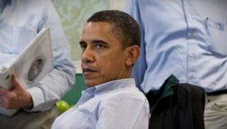 Barack Obama, wearing a light blue checkered shirt, looks intently to his left during a briefing, with two other men partially visible in the background.