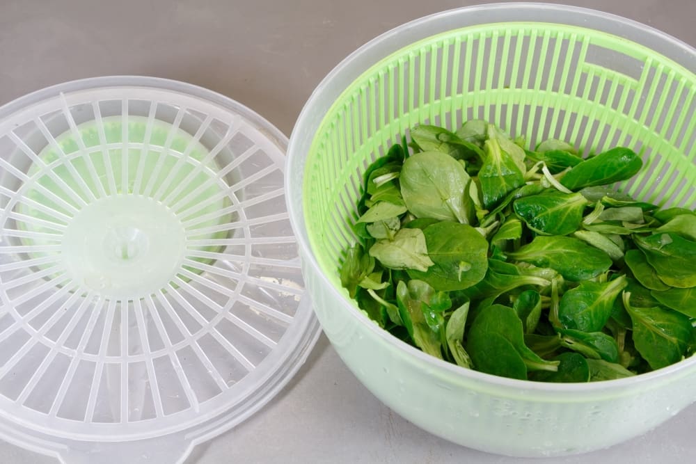 A green and white salad spinner, with fresh green lettuce leaves in its basket, and the lid resting beside it.