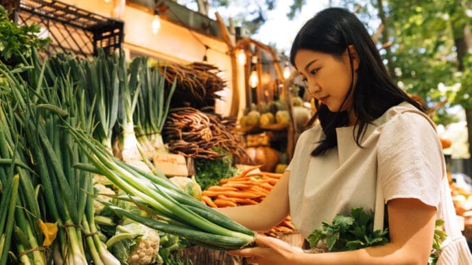 A young Asian woman with long dark hair, wearing a light-colored top, carefully selects fresh green onions at an outdoor food market with a wide variety of vegetables and warm lighting.