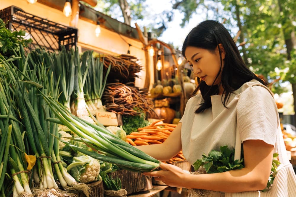 A young Asian woman with long dark hair, wearing a light-colored top, carefully selects fresh green onions at an outdoor food market with a wide variety of vegetables and warm lighting.