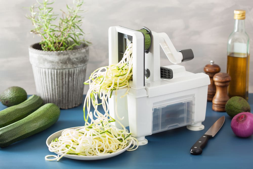 A white spiralizer actively turning raw zucchini into "zoodles," with a plate of spirals and fresh ingredients on a blue surface.