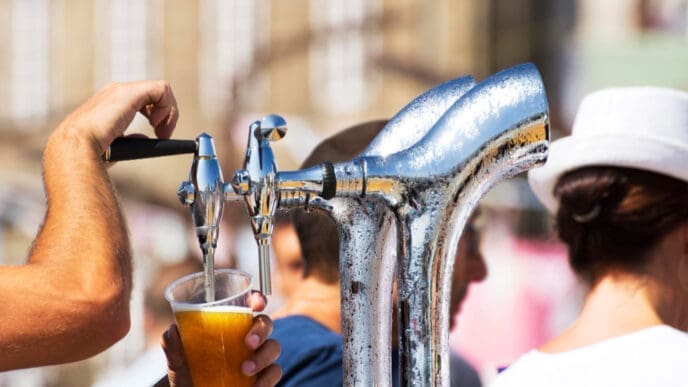 A hand pulls a lever on a shiny chrome beer tap, pouring golden beer into a clear plastic cup, with blurred people and a building in the sunny background, evoking a lively outdoor event.
